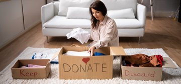 Person sorting clothes into 'Keep,' 'Donate,' and 'Discard' boxes while sitting on the floor, organizing and decluttering for a cleaner, healthier living space.