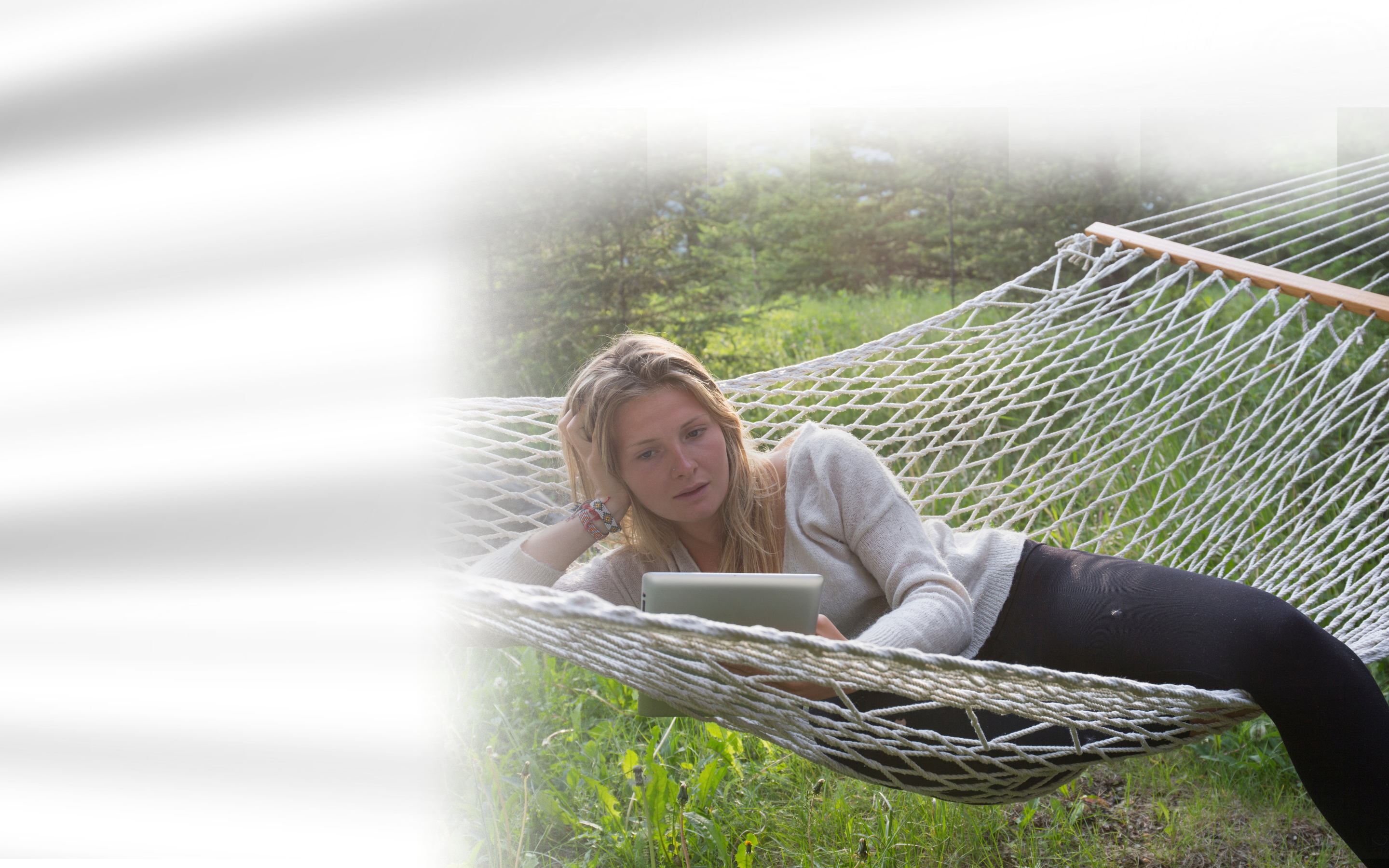 A woman with blonde hair relaxed in a rope hammock on a grassy area, wearing a sweater and dark pants while looking intently at a silver tablet held in her hands.